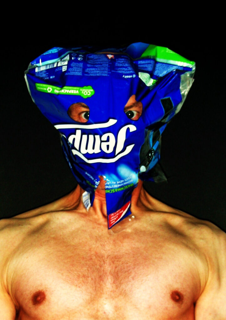Fotokunst aus der Serie „Konsumselfie“ von Künstler Nede (Peter Debusi). Das farbintensive Porträt vor tiefschwarzem Hintergrund zeigt den nackten Oberkörper des Künstlers frontal zum Betrachter. Sein Kopf ist vollständig von einer plastischen, blau-grünen Kunststoffmaske verhüllt, die aus einer leeren Großpackung von „Tempo“-Taschentüchern gefertigt wurde. Die Verpackung ist kopfüber aufgesetzt, sodass der ikonische, weiße Markenschriftzug „Tempo“ kopfsteht. Durch zwei präzise ausgeschnittene Sehschlitze direkt über dem Schriftzug blicken die Augen des Künstlers intensiv hervor. Am Hals verjüngt sich die Plastikmaske und gibt den Blick auf Textfragmente wie „WASCHMASCHINENFEST“ frei, während die Schulter- und Brustpartie in warmen Hauttönen ausgeleuchtet ist.
