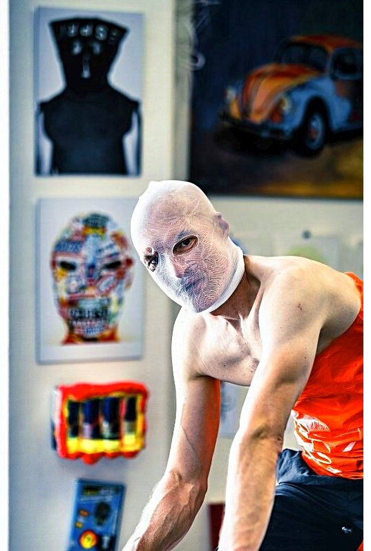 Fotokunst aus der Serie „Konsumselfie“ von Künstler Nede (Peter Debusi), fotografiert von Veit Matuschek im Rahmen der Popup-Ausstellung „illart“ in Darmstadt. Das Porträt zeigt den Künstler im Vordergrund in einer leicht gebeugten, dynamischen Pose vor einer weißen Galeriewand. Sein Kopf ist vollständig von einer eng anliegenden, weißen Nylonstrumpfhose verhüllt, durch die seine Augen intensiv blicken. Er hat einen nackten Oberkörper, während ein leuchtend orangefarbenes Element an seiner Seite zu sehen ist. Im Hintergrund hängen mehrere seiner konsumkritischen Exponate an der Wand: ganz oben eine dunkle Silhouette mit der kopfstehenden Aufschrift „ESPRIT“, darunter ein farbenfrohes Porträt-Exponat aus bunten Verpackungen sowie ein plastisches, rotes Objekt. Rechts oben im Hintergrund ist zudem ein fremdes Gemälde eines bunten VW Käfers zu erkennen.