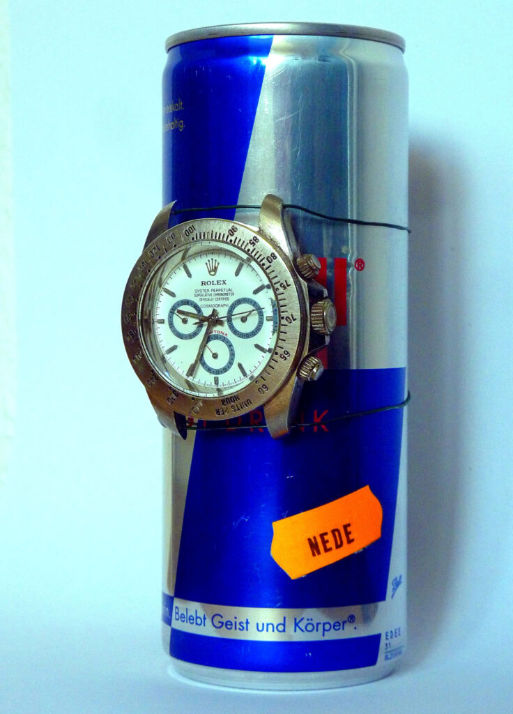 Plastische Kunst von Künstler Nede (Peter Debusi) mit dem Titel „Zeitbombe“. Das Objekt vor hellem Hintergrund besteht aus einer ungeöffneten, schlanken Red-Bull-Dose im typischen blau-silbernen Design. Auf der Vorderseite der Dose ist das Gehäuse einer goldenen Rolex-Daytona-Armbanduhr (Chronograph) mit weißem Zifferblatt montiert. Die Uhr ist mit zwei feinen, schwarzen Drähten, die horizontal um den Dosenkörper verlaufen, straff fixiert, was an die Konstruktion eines Sprengsatzes erinnert. Unten rechts auf der Dose klebt schräg ein leuchtend orangefarbenes Preisetikett mit deinem Künstlernamen „NEDE“. Am unteren Rand ist der Slogan „Belebt Geist und Körper.®“ gut lesbar.