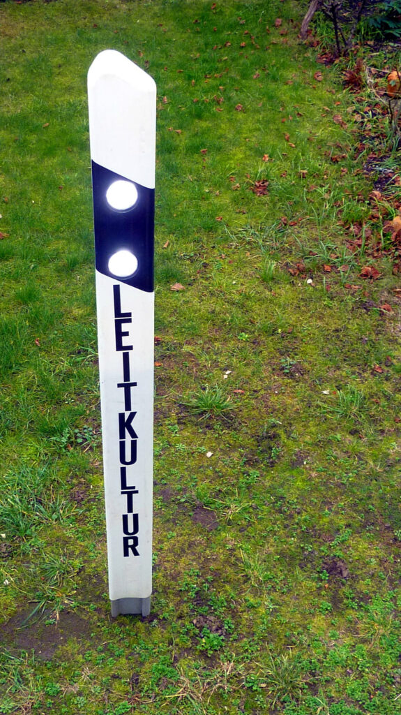 Konzeptkunst von Künstler Nede (Peter Debusi) zum Thema „Leitkultur“. Die Fotografie zeigt ein Kunstobjekt im Außenbereich, das auf einer Rasenfläche mit Moos und herbstlichen Blättern aufgestellt ist. Das Objekt ist ein klassischer, weißer Straßenleitpfosten aus Kunststoff mit einer abgeschrägten Oberseite. Im oberen, schwarzen Schrägbalken des Pfostens sind zwei kreisrunde, hell leuchtende oder reflektierende weiße Lichter integriert. Vertikal untereinander verläuft auf der weißen Vorderseite des Pfostens das Wort „LEITKULTUR“ in serifenlosen, schwarzen Großbuchstaben.