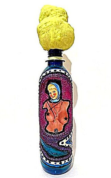 Plastische Kunst von Künstler Nede (Peter Debusi) mit dem Titel „Flaschenpost“. Das Objekt vor weißem Hintergrund erinnert an einen Molotow-Cocktail und thematisiert Träume, Erotik und Werbung. Es besteht aus einer aufrecht stehenden, blau glänzenden Glasflasche, aus deren Flaschenhals eine große, quillende Masse aus hellgelbem Bauschaum wie eine Rauchwolke emporragt. Der Flaschenkörper ist kunstvoll verziert: Im Zentrum befindet sich das farbige Motiv einer lächelnden, nackten Pin-up-Frau mit blauem Kopftuch und einer Kette um den Hals, deren Brüste mit schwarzen Sternen versehen sind. Das Frauenporträt ist eingebettet in eine glitzernde, lila-pinkfarbene Fläche, die von einem schwarz-weiß gepunkteten, wellenförmigen Rahmen und blauem Glitzer im unteren Bereich umschlossen wird.