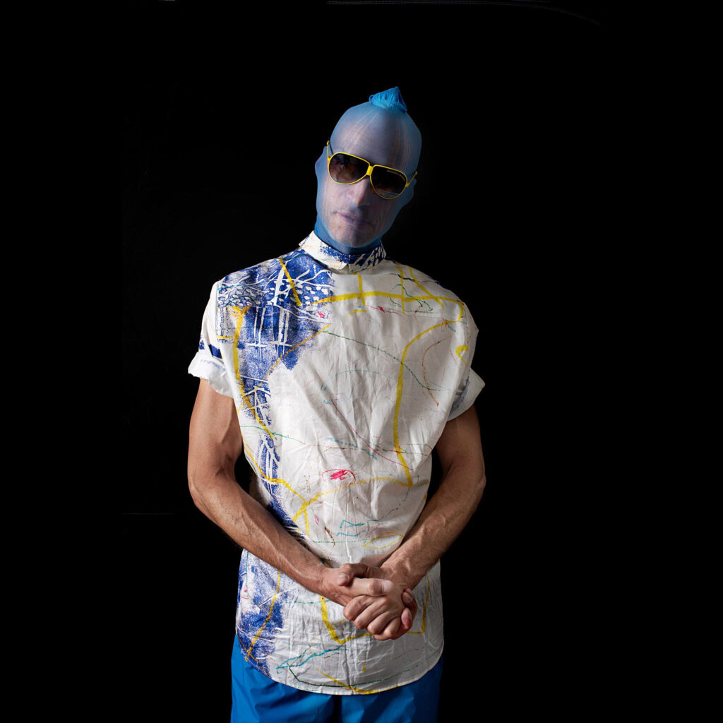 Künstler-Porträt von NEDE (Peter Debusi) als Model in einem GERMENS Artfashion Designerhemd von Heinz W. Lotz, fotografiert von Richard Byrdy. Das Bild zeigt eine männliche Person vor tiefschwarzem Hintergrund in der Frontalansicht. Sie trägt ein kunstvoll gemustertes, weißes Kurzarmhemd mit blauen und gelben Pinselstrichen sowie abstrakten Linien. Über den Kopf und das Gesicht ist eine eng anliegende, transparente blaue Maske oder ein Schleier gezogen, der oben spitz zuläuft. Dazu trägt die Person eine auffällige Sonnenbrille mit gelbem Rahmen und dunklen Gläsern. Die Hände sind vor dem Bauch ineinandergelegt.