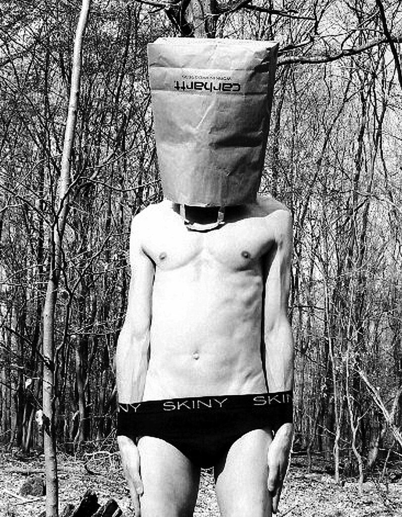 Konsumselfie des Künstlers NEDE (Peter Debusi) zum Thema Konsum, Markenidentität und Entfremdung, bei dem der gesamte Kopf in einer braunen Papiertüte der Marke Carhartt steckt. Das Schwarz-Weiß-Foto zeigt den nackten Oberkörper des Künstlers von vorn vor einer dichten Waldkulisse im Hintergrund. Der umgedrehte Schriftzug „carhartt“ zieht sich gut lesbar über den oberen Teil der Papiermaske. Am Körper trägt der Künstler eine dunkle Unterhose mit der deutlich sichtbaren Aufschrift „SKINY“ auf dem elastischen Bund. Das Werk thematisiert visuell die Anonymisierung des Individuums und den allgegenwärtigen Markenzwang.