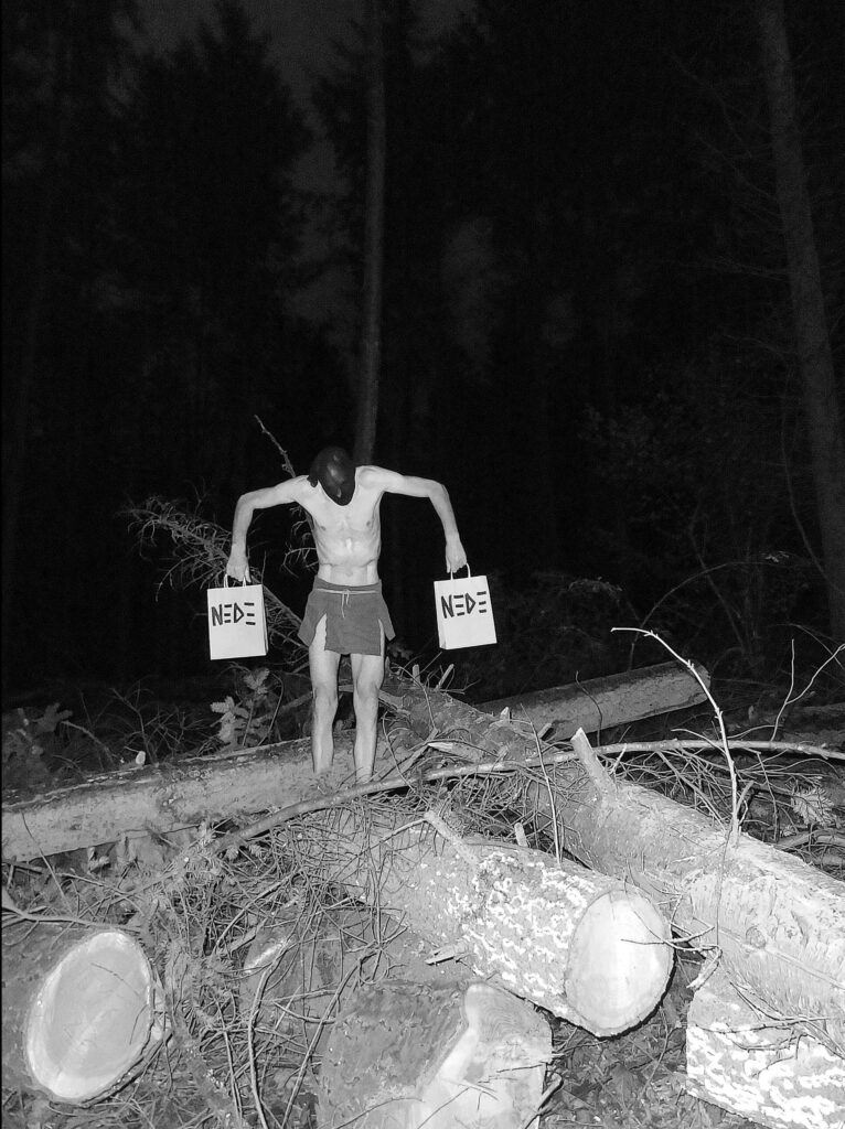 Fotokunst des Künstlers NEDE (Peter Debusi) zum Thema Konsumwahn, Zerstörung und Markenidentität mit dem Titel „Black Friday Warmup“. Das kontrastreiche Schwarz-Weiß-Foto zeigt eine Performance im Wald, die an eine Rambo-Szene erinnert. Der Künstler steht mit nacktem Oberkörper, einer dunklen Gesichtsmaske und einem improvisierten Rock inmitten von gefällten Baumstämmen und grobem Geäst einer Rodung. In jeder Hand trägt er mit angewinkelten Armen eine weiße Papiertüte mit dem großen, schwarzen NEDE-Logo. Das Werk thematisiert visuell die Brutalität des modernen Konsums und den Kampf des Individuums in einer zerstörten Umwelt.